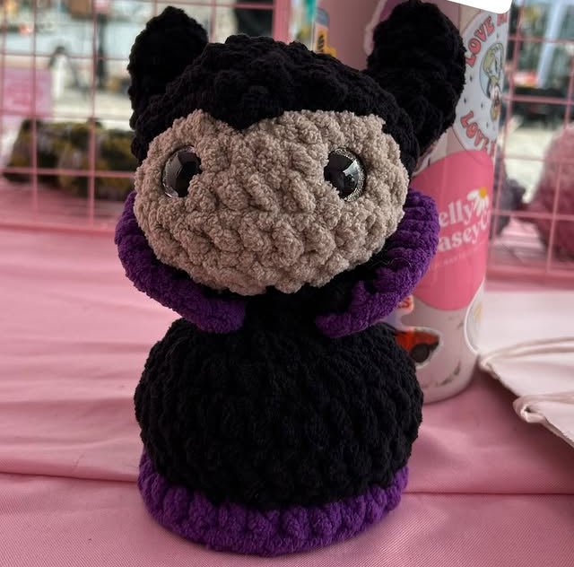 Crocheted character with black, purple, and gray colors on a pink surface.